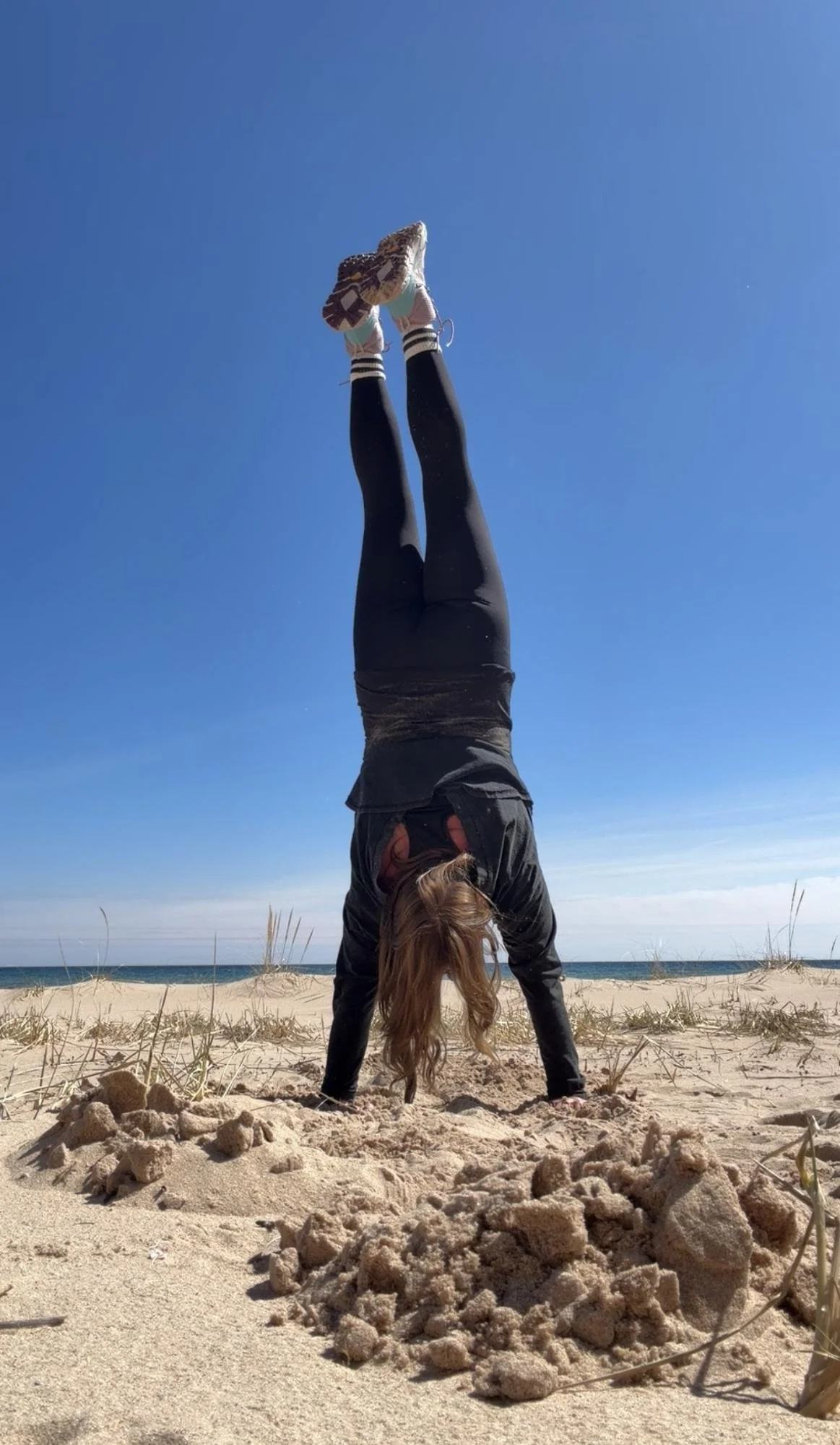 A person doing a handstand on a sandy beach with clear blue sky and ocean in the background. They are wearing black leggings, sneakers, and a dark jacket, with long hair hanging down.