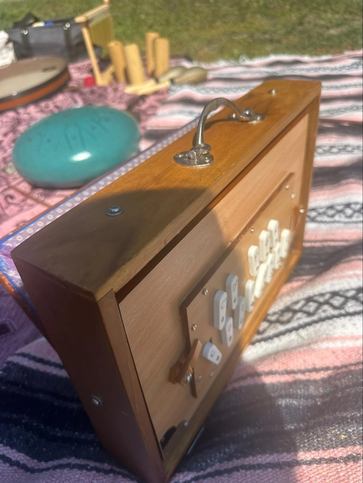 A vintage wooden cabinet with white knobs, placed on a patterned blanket on the grass, displaying a blue oval-shaped musical instrument, possibly a steel drum, in the foreground. In the background, there are various musical instruments and wooden per