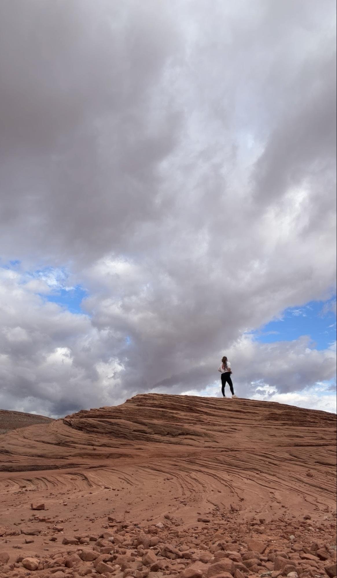 A person standing on a rocky desert landscape with layered reddish-brown rocks, under a cloudy sky with patches of blue.