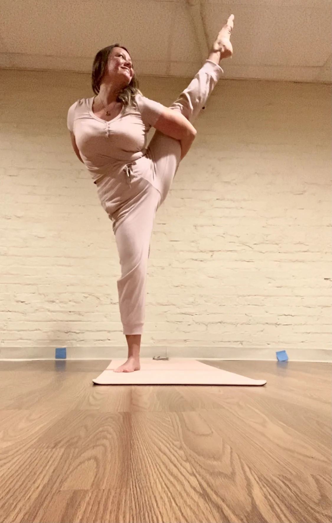 A woman in beige clothing practicing yoga indoors, standing on a white yoga mat, performing a one-legged pose with her left leg raised high behind her, holding her foot with her left hand, while her right hand rests on her waist. She is smiling and l