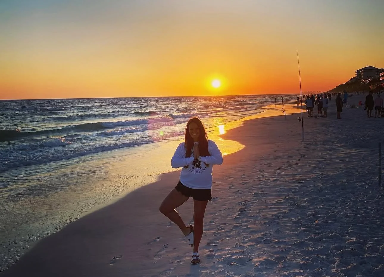 A woman practicing yoga on the beach during sunset with other people walking and relaxing in the background.