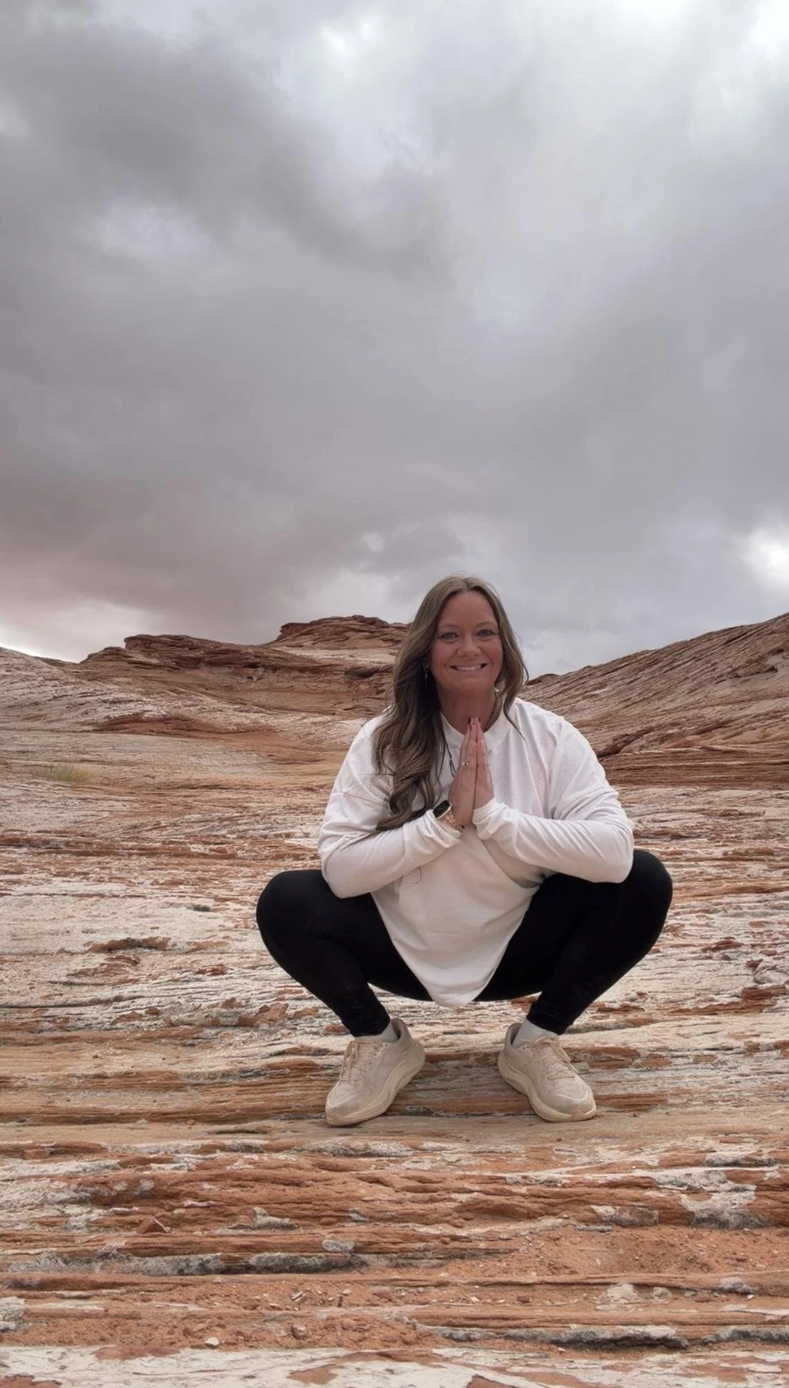 A woman practicing yoga outdoors on a rocky desert landscape, squatting with hands in prayer position, under a cloudy sky.