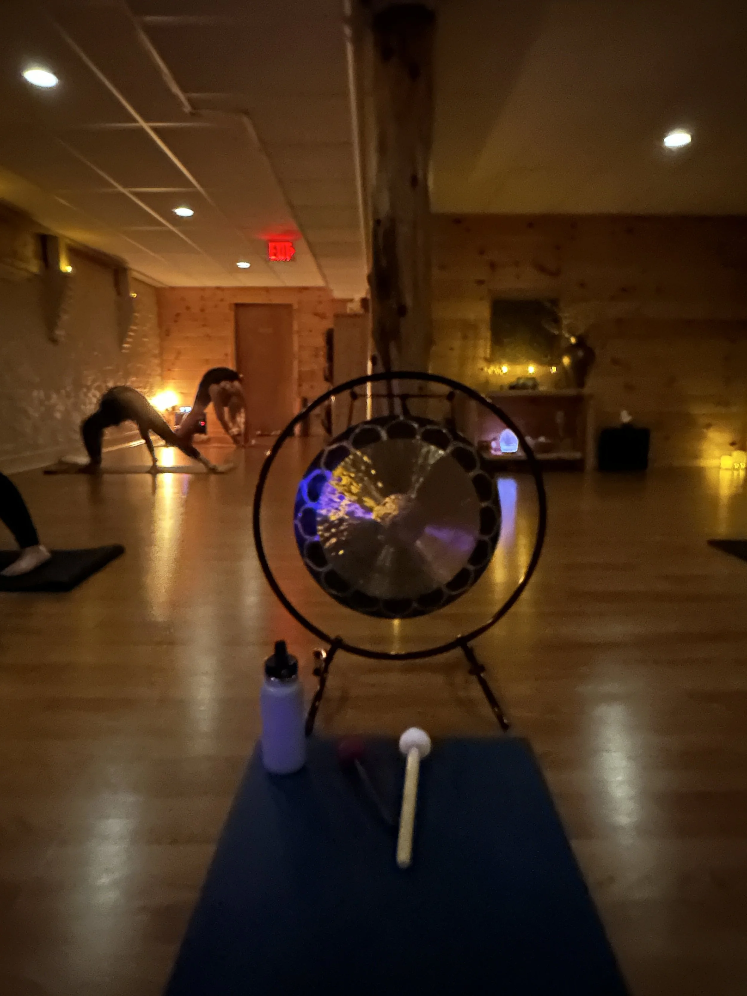 Yoga class in a dimly lit studio with a woman doing a yoga pose in the background. Yoga mats and bottles are on the floor, with the photo taken from a person's perspective sitting on a mat.