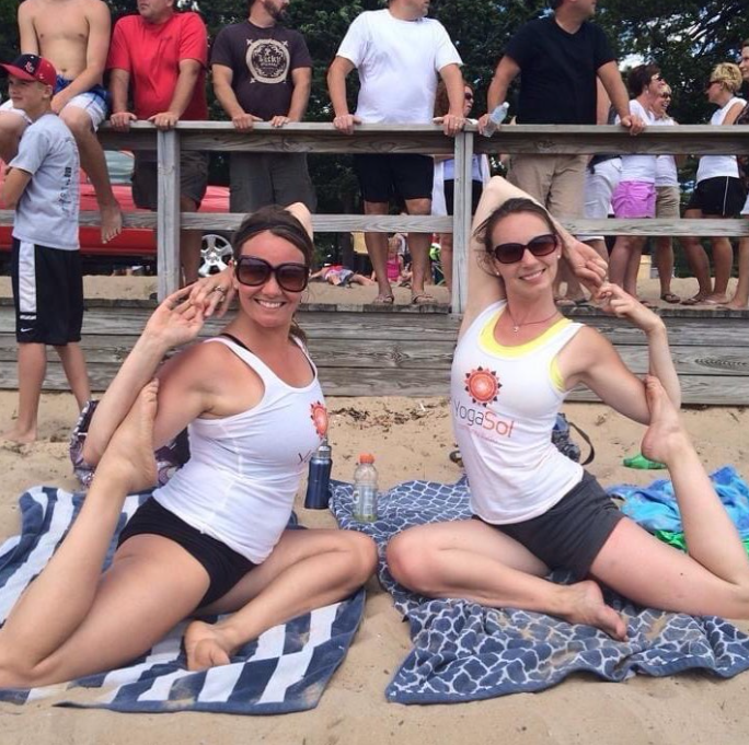 Two women in yoga tank tops and shorts sitting on beach towels on sand, posing with their legs extended and hands behind their ears, smiling. Behind them is a wooden railing with people standing, some in casual summer clothes, at what appears to be a