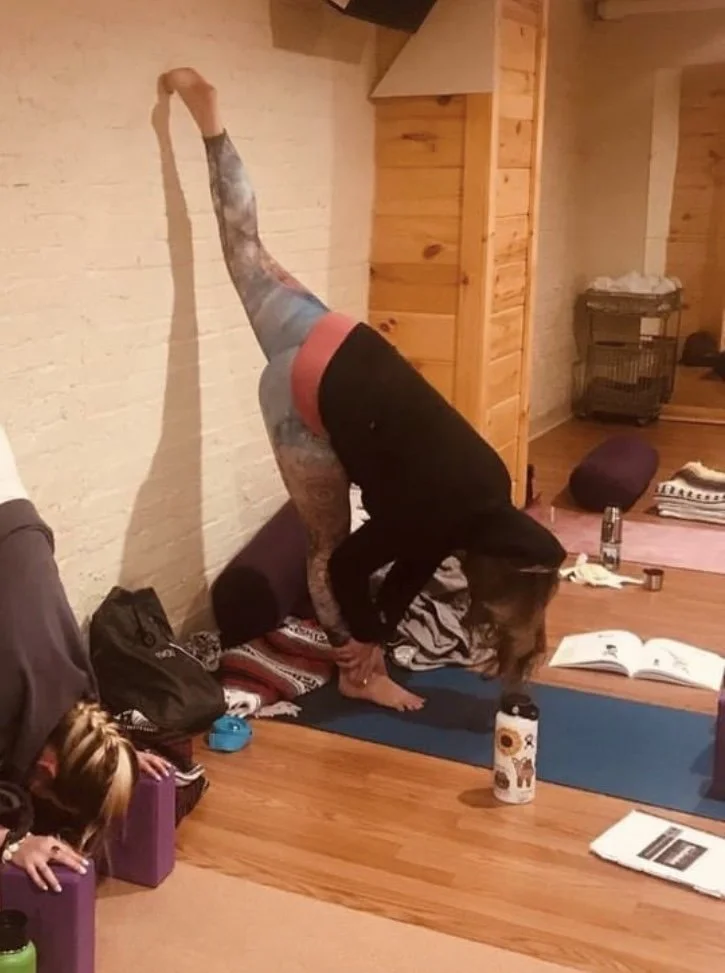 Person practicing a headstand yoga pose in a room with wood-paneled walls, surrounded by yoga mats, books, and water bottles, with a young girl watching nearby.