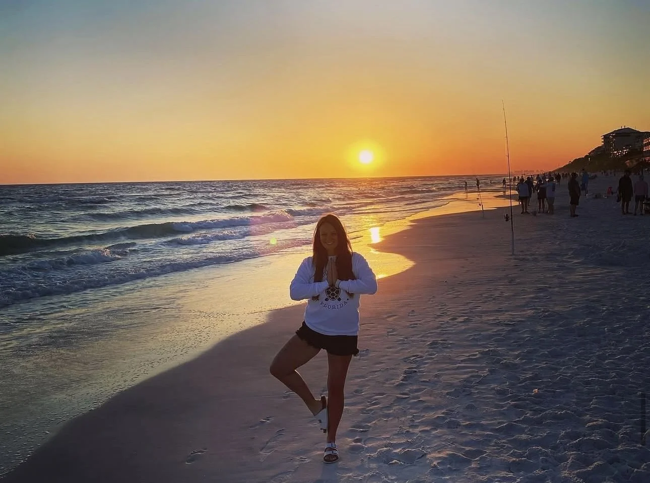 A woman practicing yoga on a sandy beach during sunset with the ocean behind her and people in the distance.