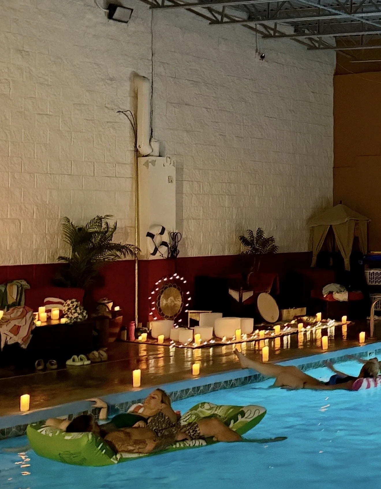 Indoor swimming pool area decorated with candles, plants, and decorative items for a gathering or event, with people relaxing on inflatable floats in the pool.