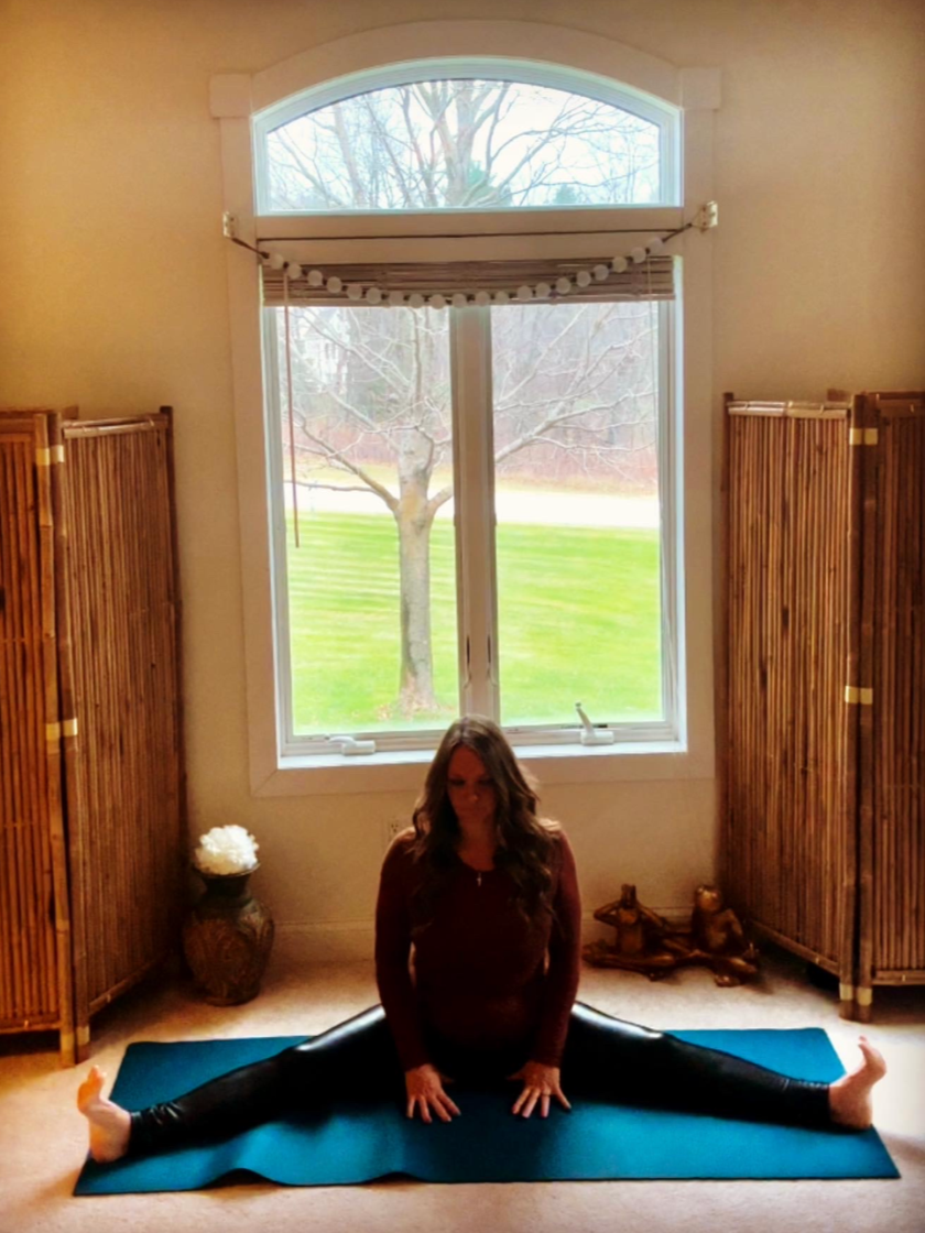 A pregnant woman in a dark red top and black leggings performing a split on a blue yoga mat indoors, with a large window showing a tree and green grass outside.
