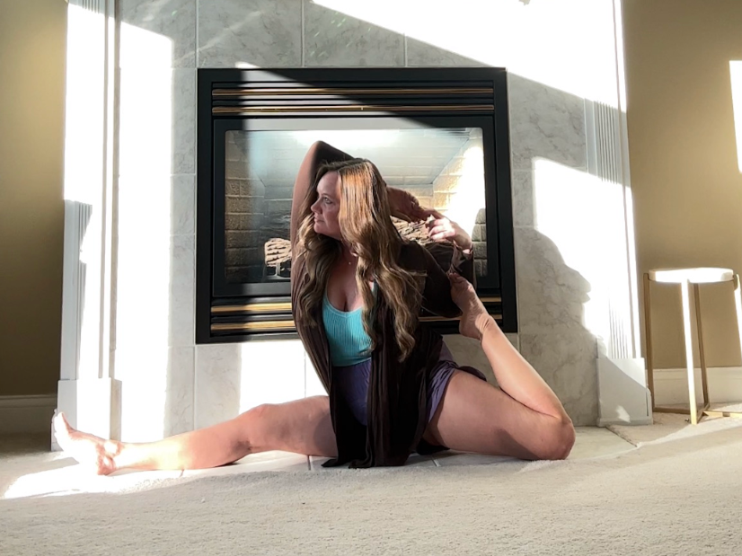 A woman practicing yoga in front of a fireplace in a living room, sitting in a split position with her arms raised and hands behind her head.