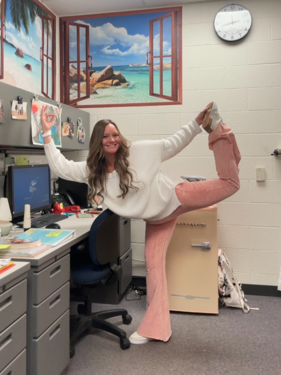A woman in an office doing a yoga pose with one leg raised behind her, holding her foot, and smiling. The office has a mural of an open window view of a beach with rocks and blue skies on the wall.
