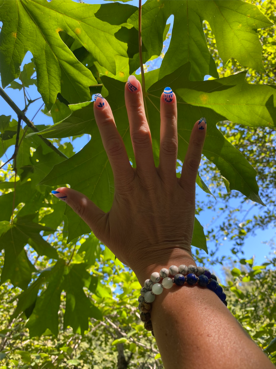Hand with three decorated painted fingernails reaching towards green maple leaves against a bright blue sky.