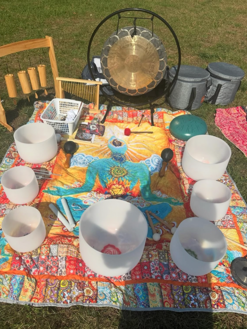 Collection of musical and meditation instruments arranged on a colorful cloth outdoors, including crystal singing bowls, a gong, cymbals, chimes, a tongue drum, mallets, and a zen garden rake.