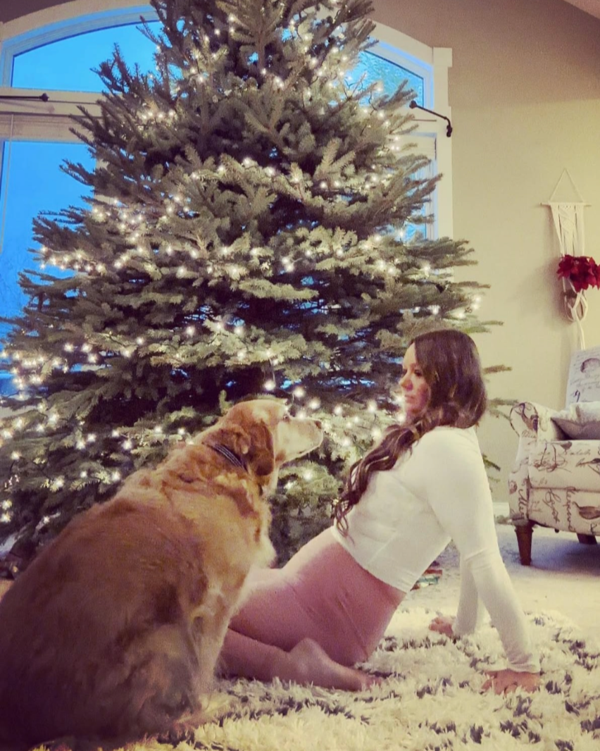 A woman sitting on a fluffy rug in front of a decorated Christmas tree with lights, with a golden retriever sitting facing her indoors during winter.