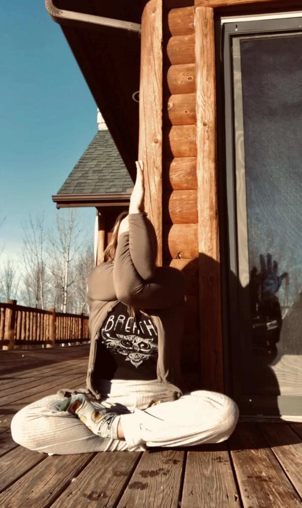 Person practicing yoga on a wooden deck outside a log cabin, sitting cross-legged with a focused pose, reflected in a large glass door, sunny day with clear sky.