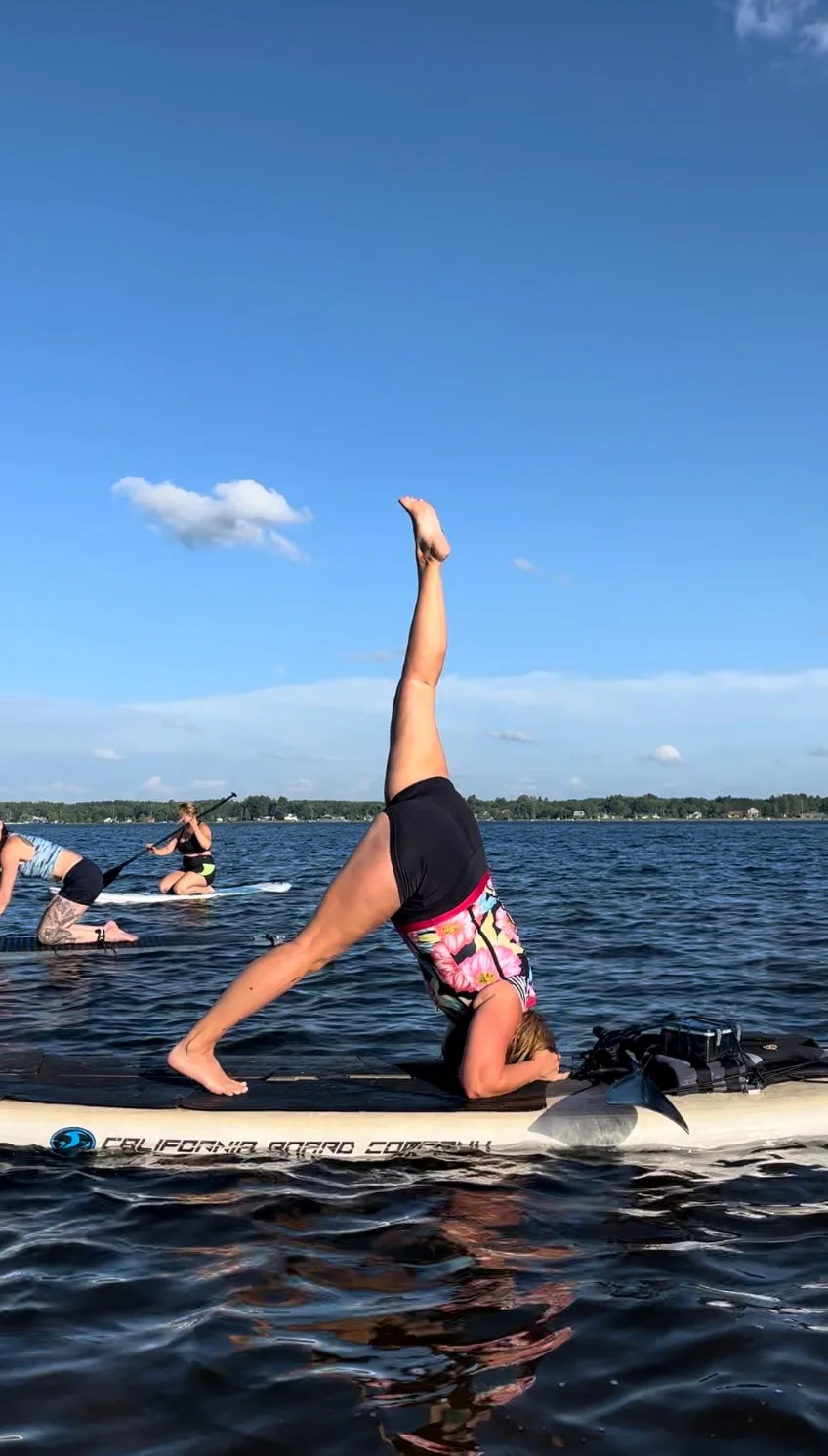 A woman practicing yoga in a headstand on a paddleboard on a lake, with other paddleboarders in the background under a blue sky with some clouds.