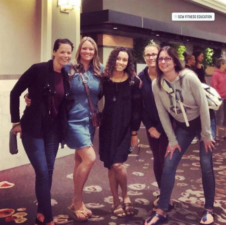 A group of five women standing together in a hotel lobby, smiling for the photo. They are dressed casually, with one woman wearing a black dress, and others in jeans and hoodies or denim shorts. The background shows some other people and greenery.