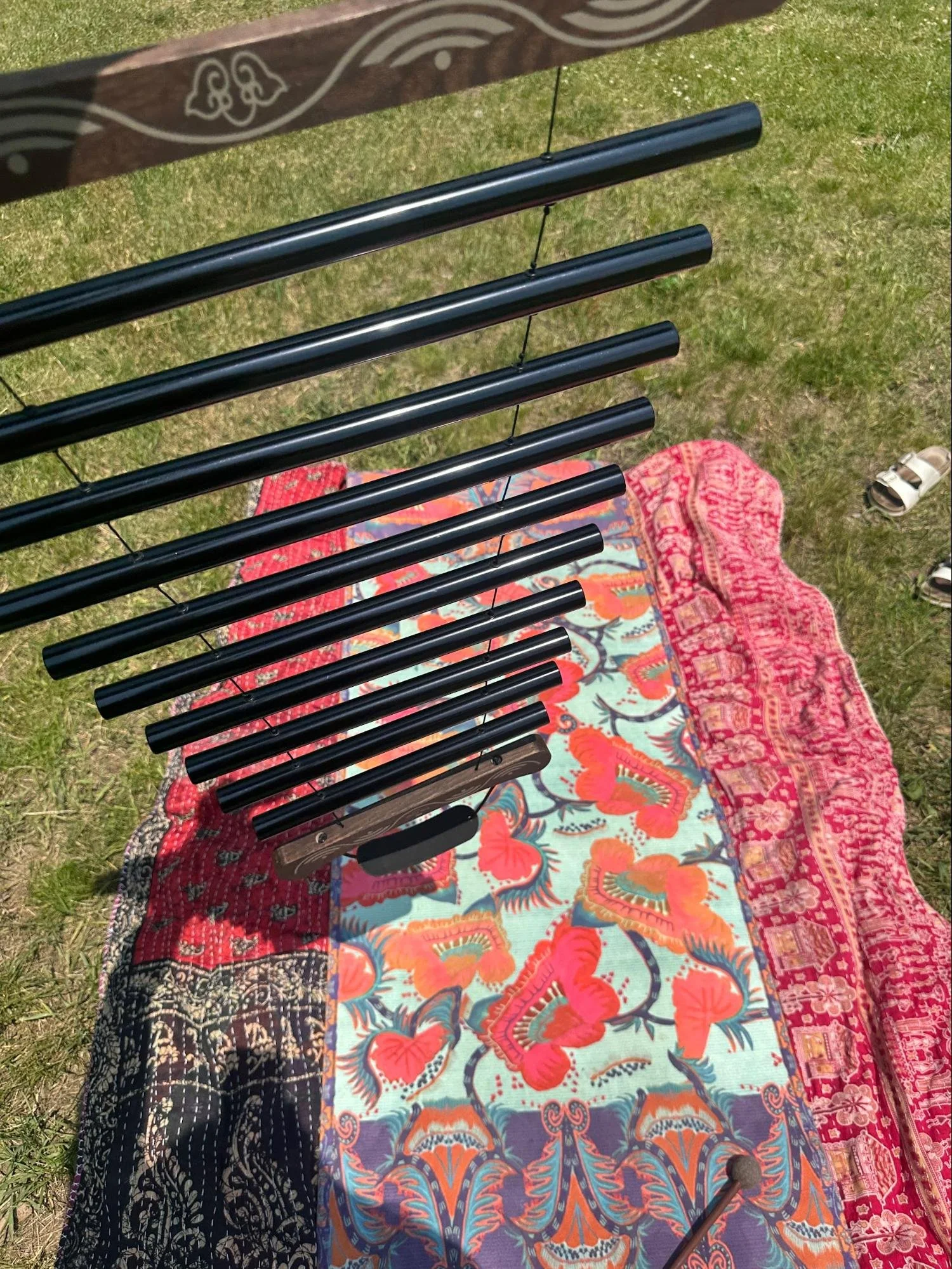 An outdoor stringed musical instrument with black tubes, possibly a wind chime, hanging above a colorful, patterned rug on the grass with sandals nearby.