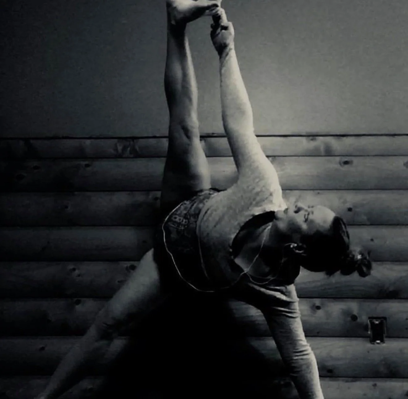 A person doing a handstand against a wooden wall, with one leg extended upward and the other bent down. The person is wearing a t-shirt, shorts, headphones, and sunglasses.