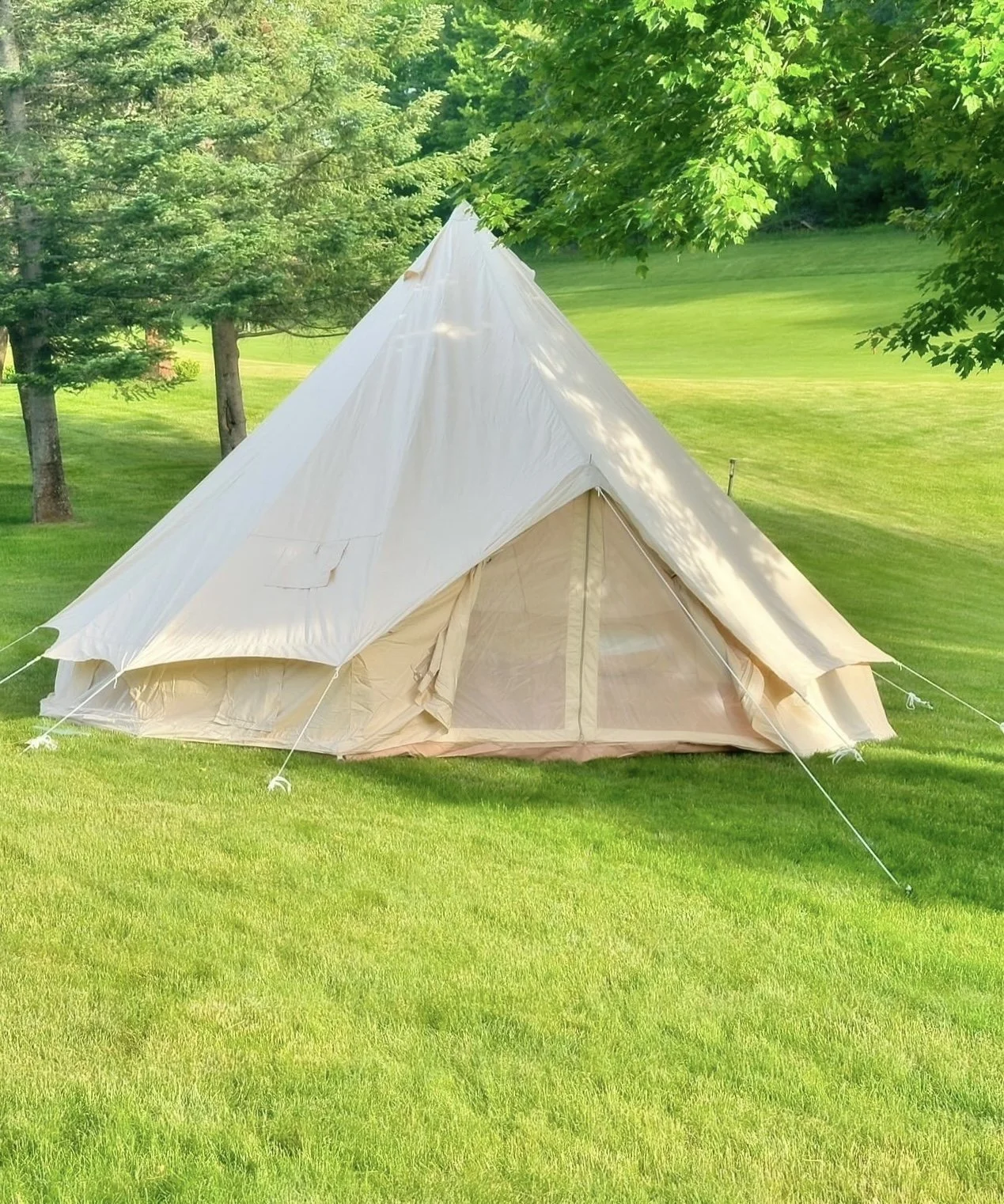 A white canvas bell tent pitched on a lush green lawn, surrounded by trees.