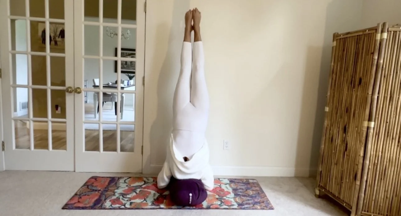 A person practicing yoga in a handstand position against a wall in a living room, with a colorful rug underneath.