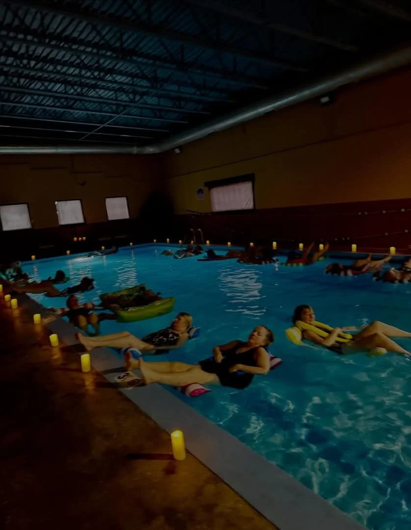 Indoor swimming pool with people relaxing on lounge chairs or floating on inflatable tubes, illuminated by candle-like lights around the pool edge, with windows and a high ceiling in the background.