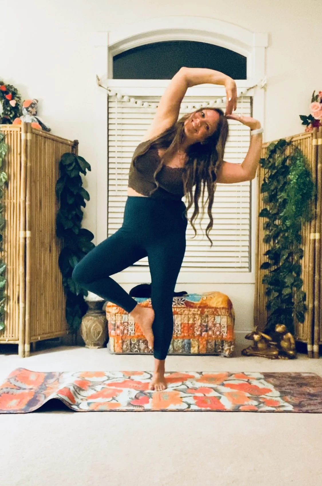 Woman practicing yoga indoors on a floral mat, balancing on one foot with arms raised in a circular shape, surrounded by plants and decorative items.