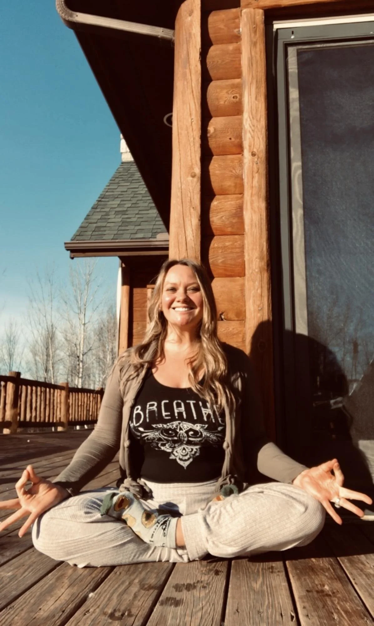 A woman in a black T-shirt with a skull design and the word 'BREATHE' on it, sitting cross-legged on a wooden deck, practicing meditation outdoors in front of a log cabin under a clear blue sky. She is smiling and has long, wavy hair.
