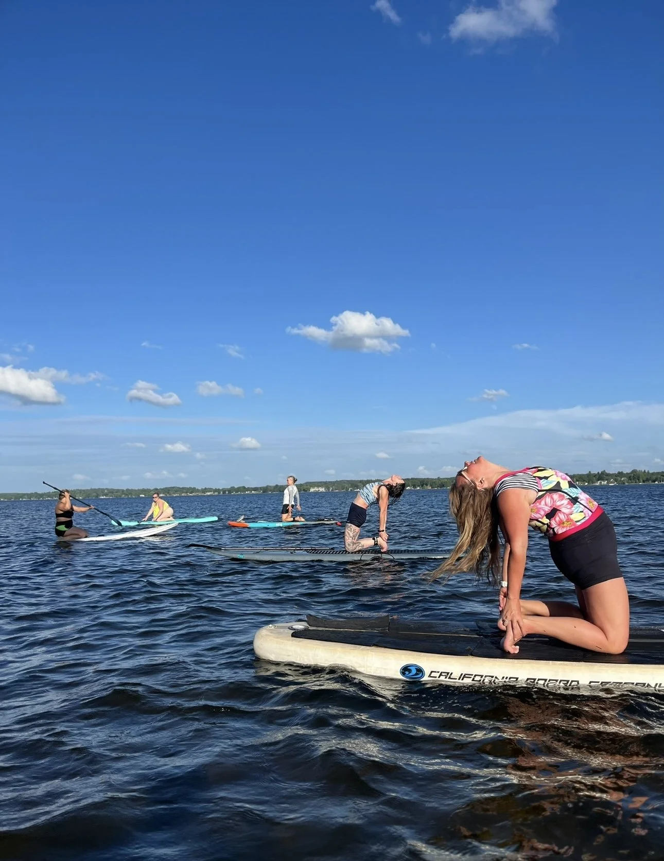 Group of people enjoying a sunny day on the water with paddleboards and a surfboard, some practicing yoga or stretching.