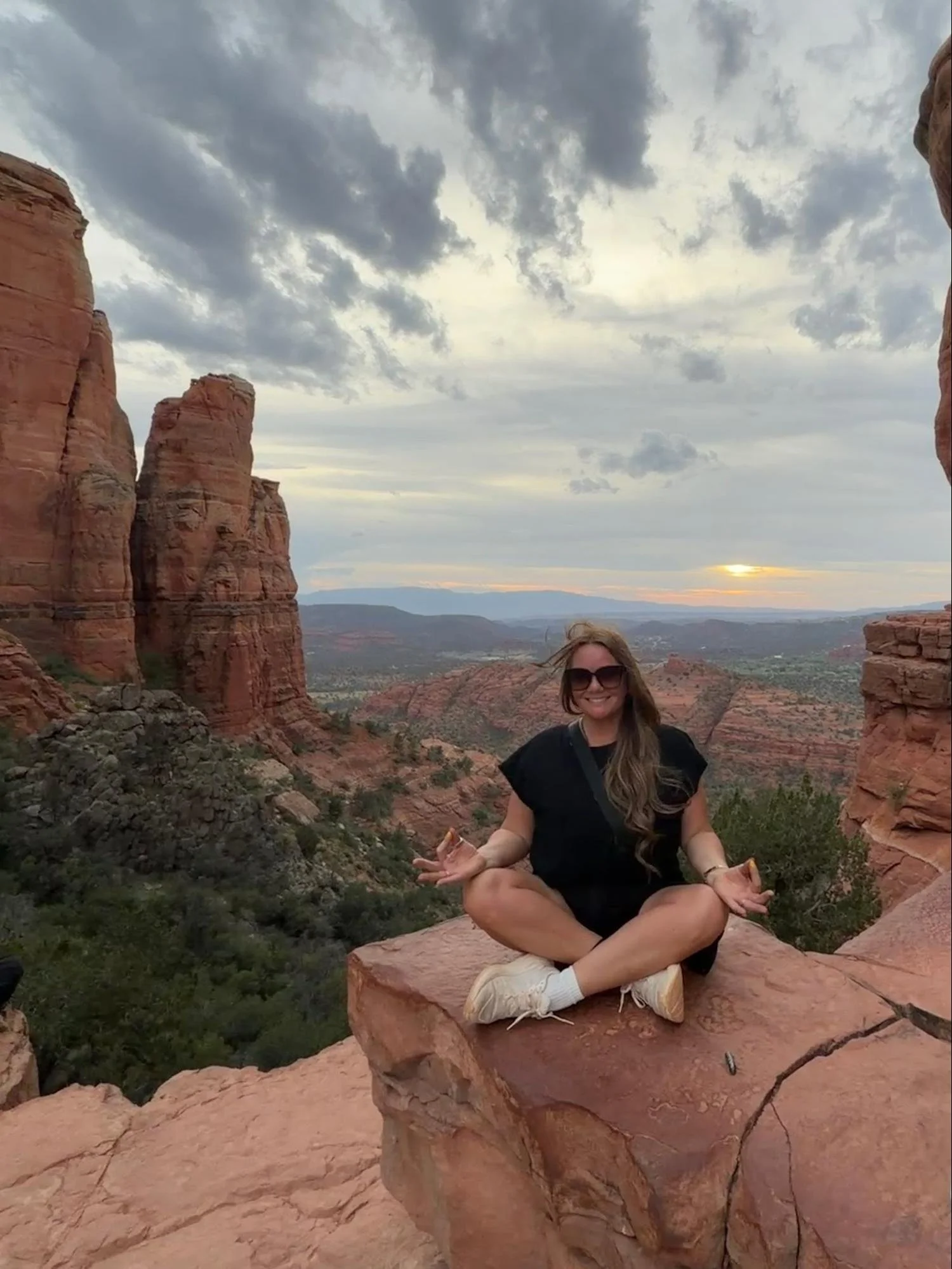 A woman sitting cross-legged on a rock ledge, meditating with a scenic view of red rock formations and a cloudy sky in the background.