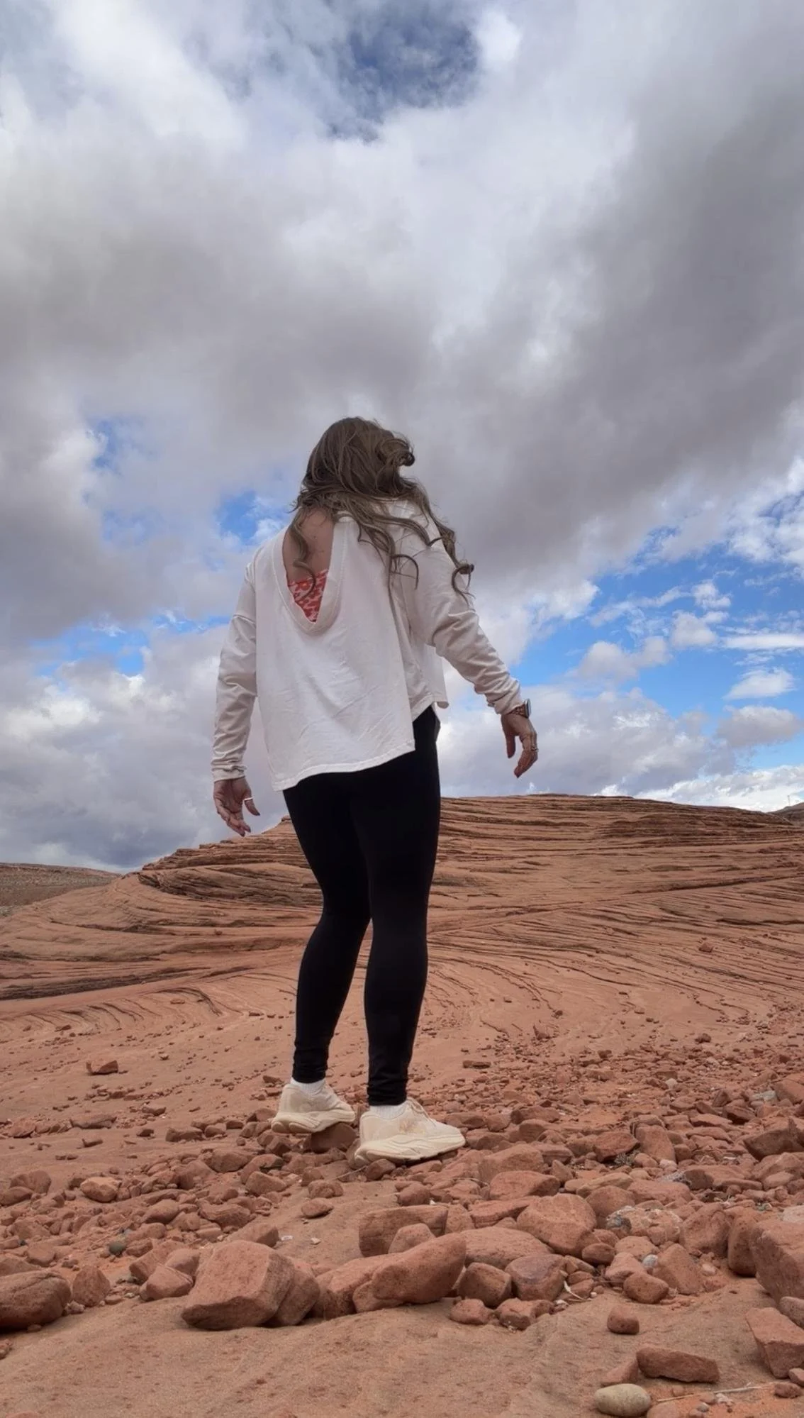 A woman with long hair, wearing a white long-sleeve shirt with holes on the sides, black leggings, and white sneakers, walking on a rocky desert landscape under a cloudy sky.