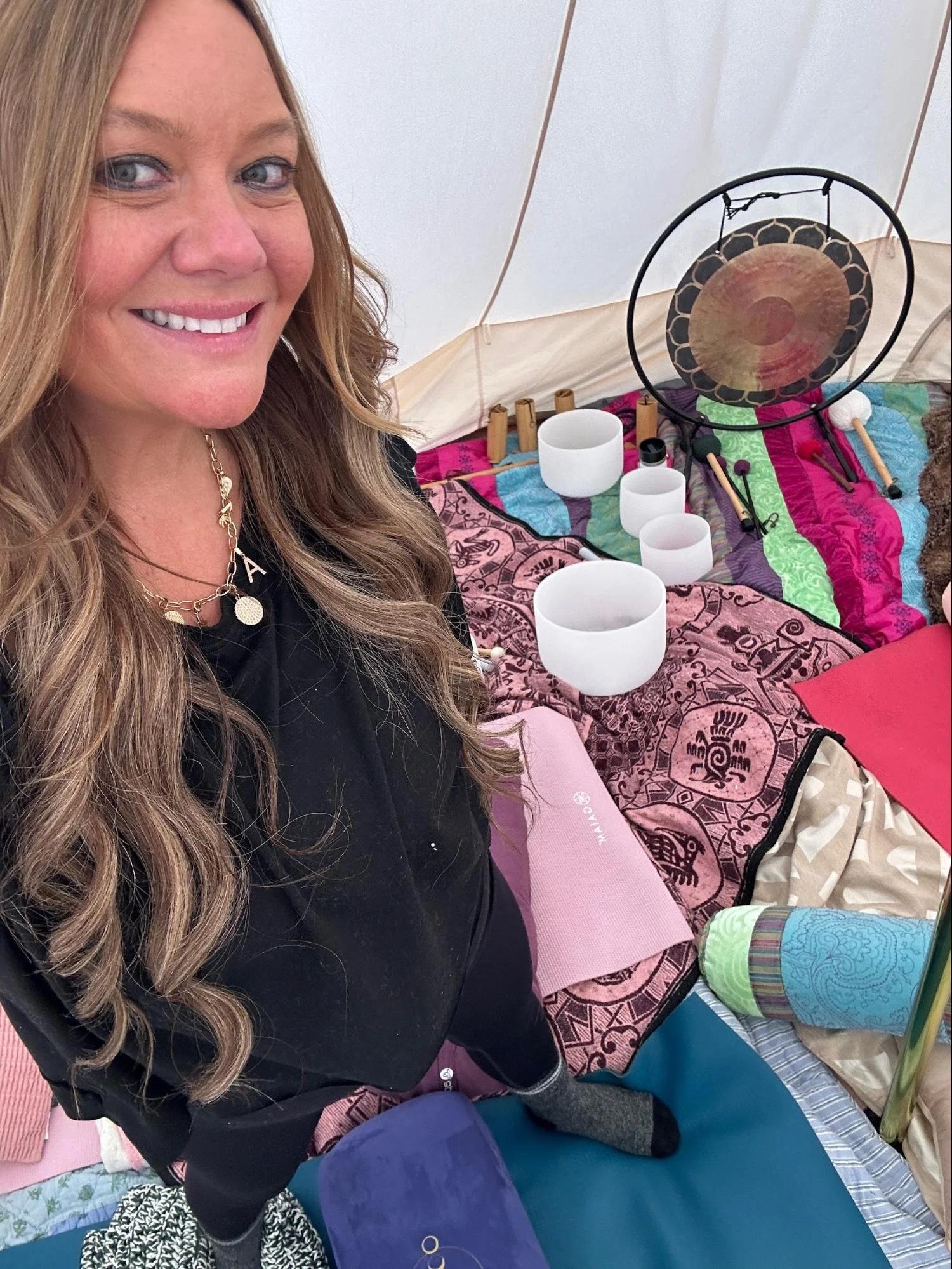Woman with long wavy hair smiling, sitting inside a tent decorated with colorful fabric and holding a drumstick, surrounded by singing bowls and musical instruments on the floor.