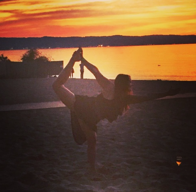 A person performs a yoga pose on a beach at sunset, with water and a distant silhouette of trees and people in the background.