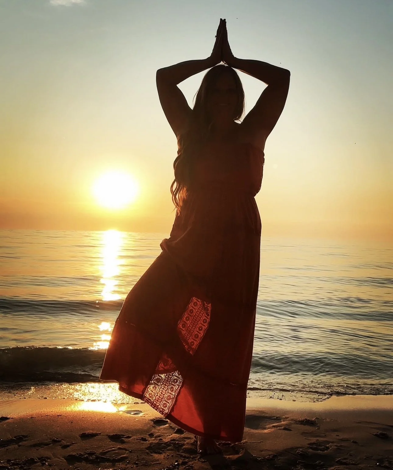 Silhouette of a woman standing on the beach with the ocean and sunset in the background, hands pressed together above her head in yoga pose.