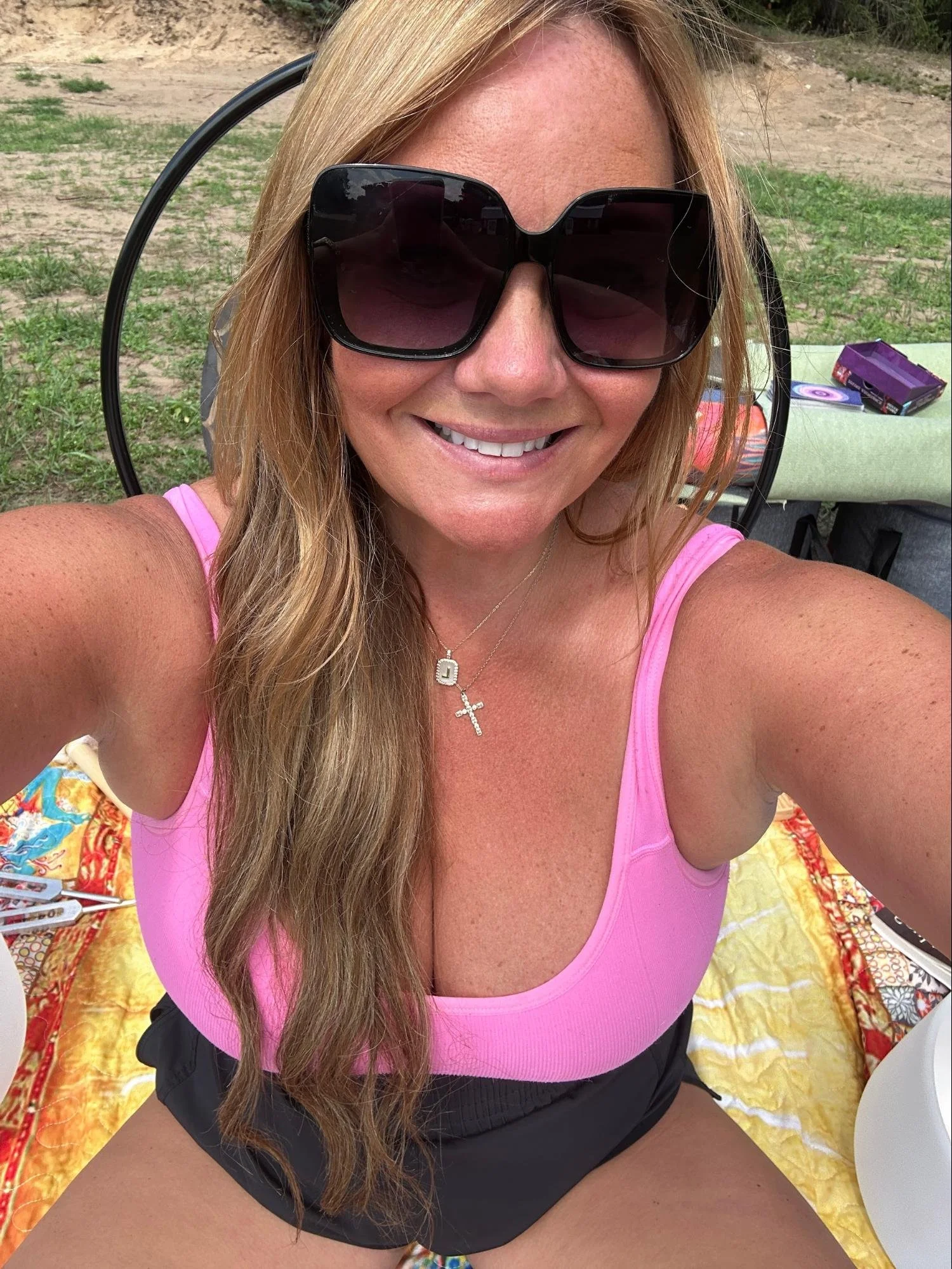 A woman taking a selfie outdoors, wearing large sunglasses and a pink tank top, sitting on a yellow patterned blanket with items like a raffle ticket and a purple box nearby.