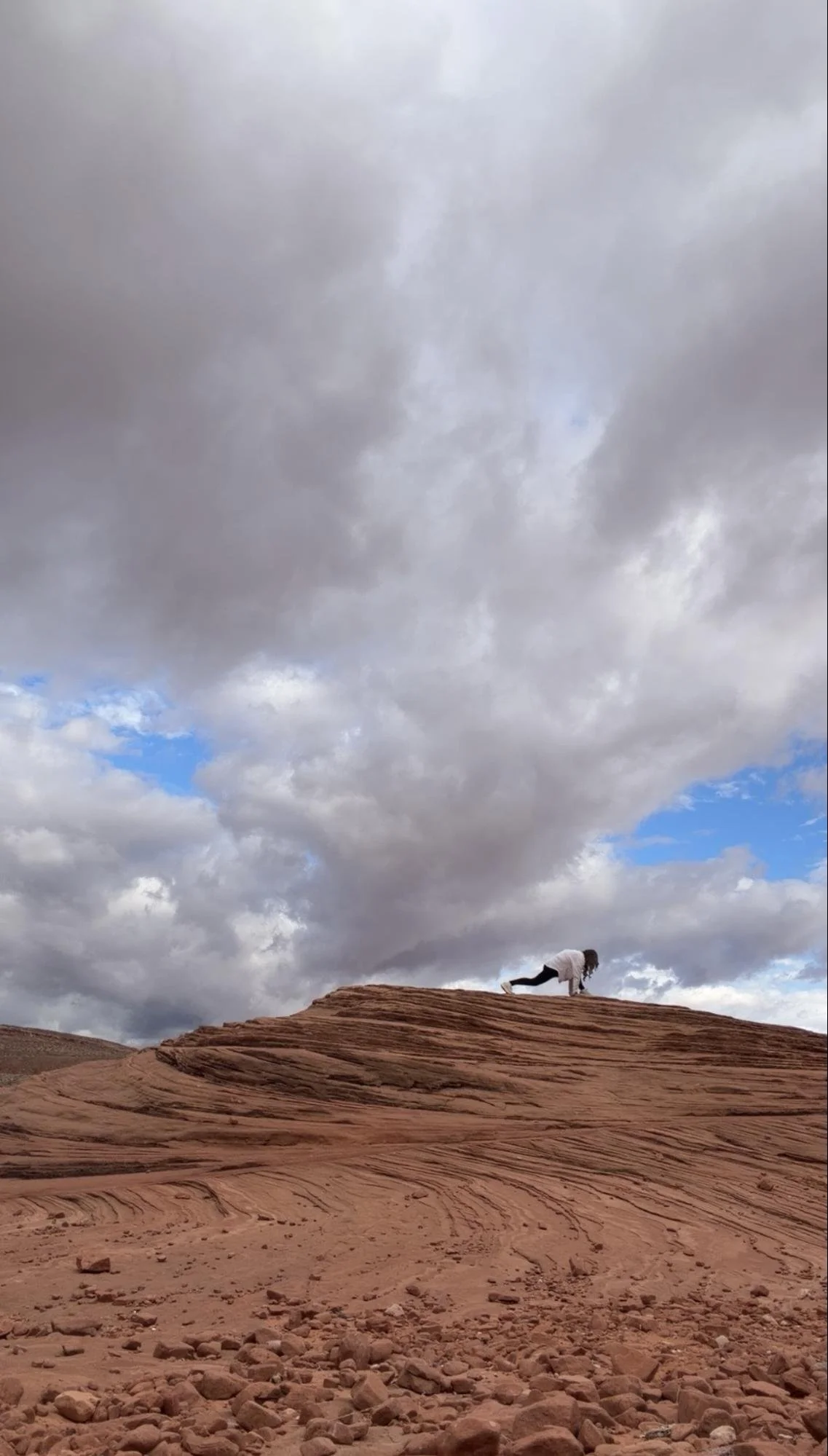 A person is doing a yoga pose on a rocky desert landscape with layered red rock formations under a cloudy sky with some patches of blue.