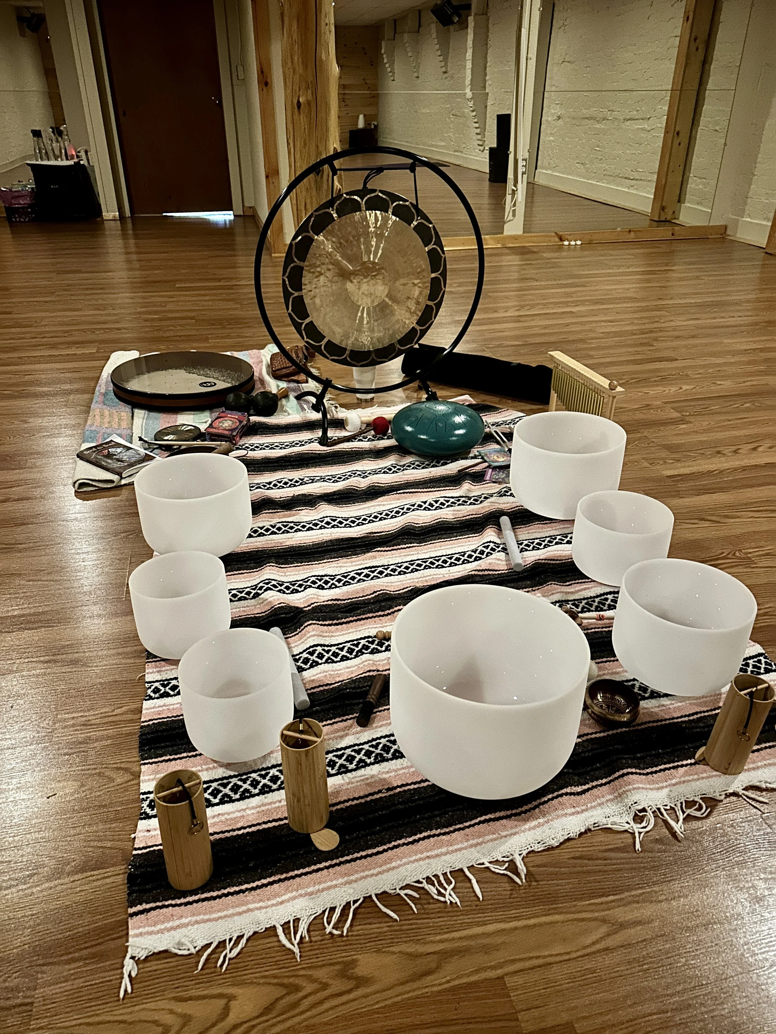 A collection of musical instruments arranged on a woven rug on a wooden floor indoors. The setup includes crystal singing bowls, bamboo percussion tubes, a gong, a tuning fork, and small mallets, indicating a sound healing or meditation session.