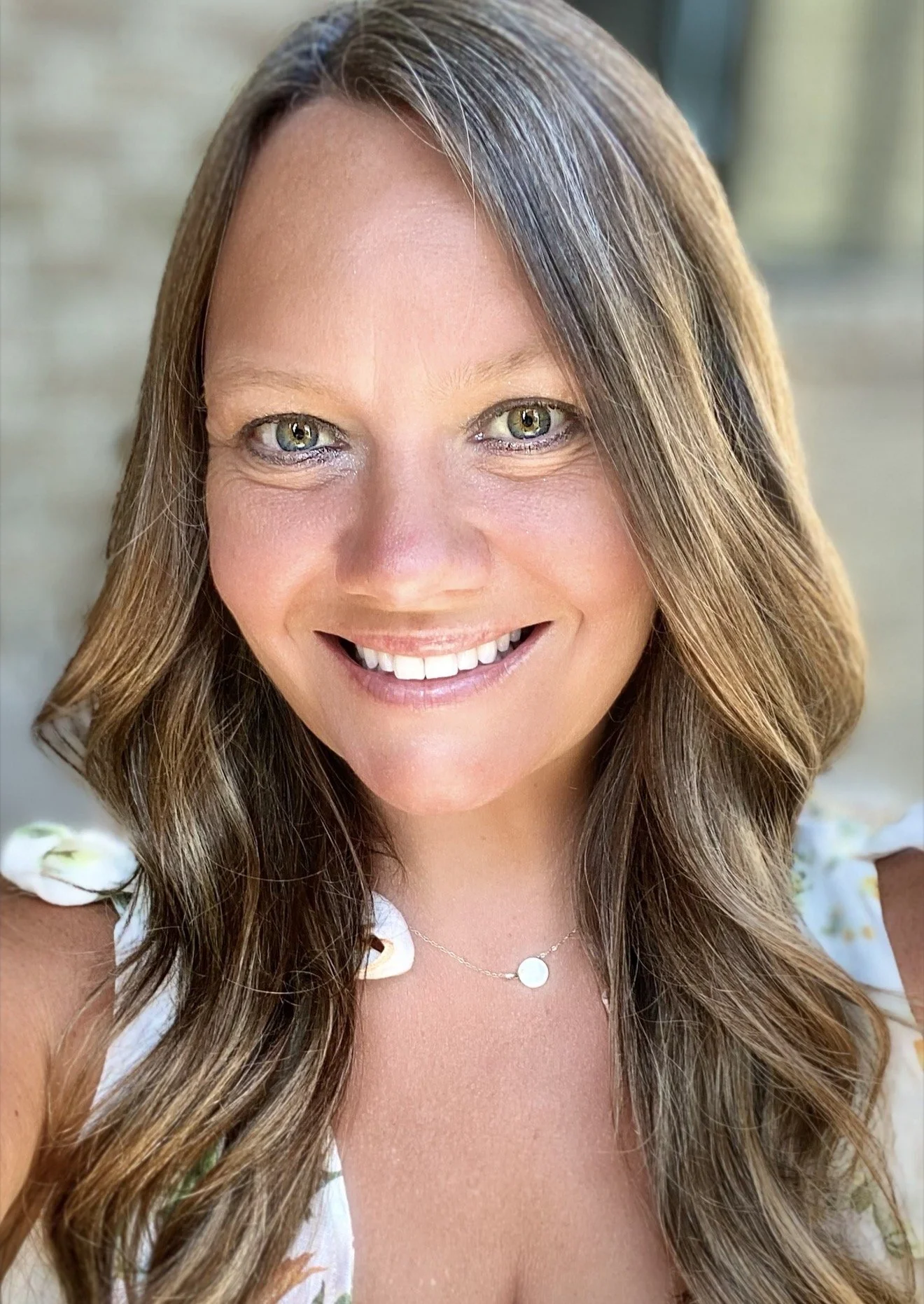 A close-up portrait of a woman with long, wavy brown hair and light eye color, smiling with white teeth, wearing a delicate necklace and a floral top, on a blurred neutral background.