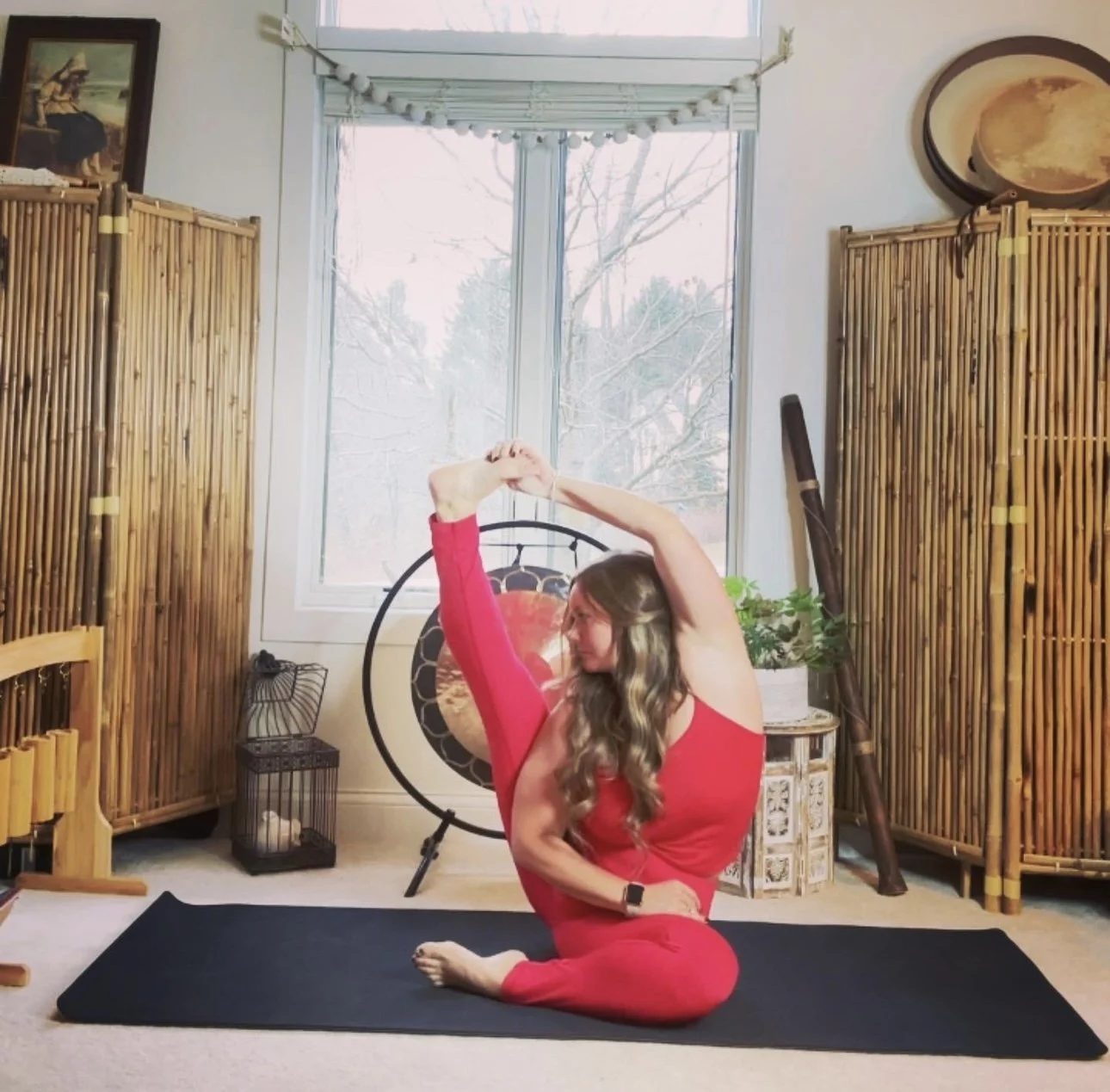 A woman in a red workout outfit doing a seated side stretch on a yoga mat in a room with bamboo room dividers, a large window, and hanging decor.
