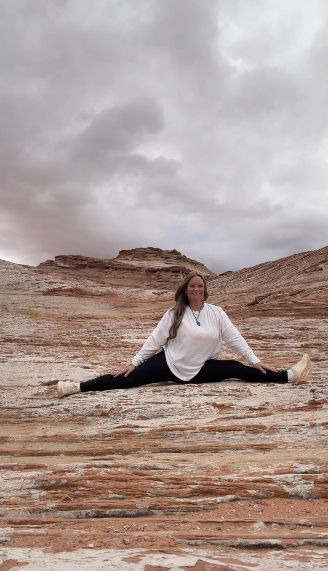 Woman sitting in a split pose outdoors on rocky terrain, with a cloudy sky in the background.
