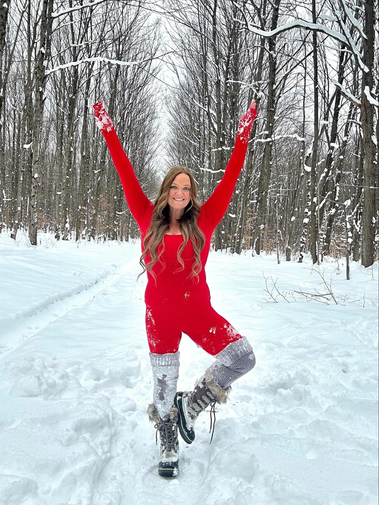 Woman in a red dress and gray knit knee-high socks posing with arms raised in a snowy forest.