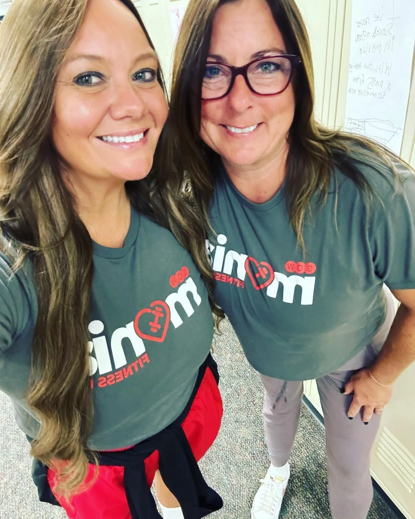Two smiling women wearing matching gray t-shirts with red and white text, standing indoors in front of a wall with a whiteboard and yellow vault door.