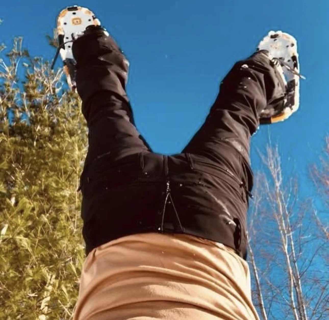 Person lying on their back looking up at the clear blue sky, wearing outdoor clothing and holding a camera, with trees in the background.