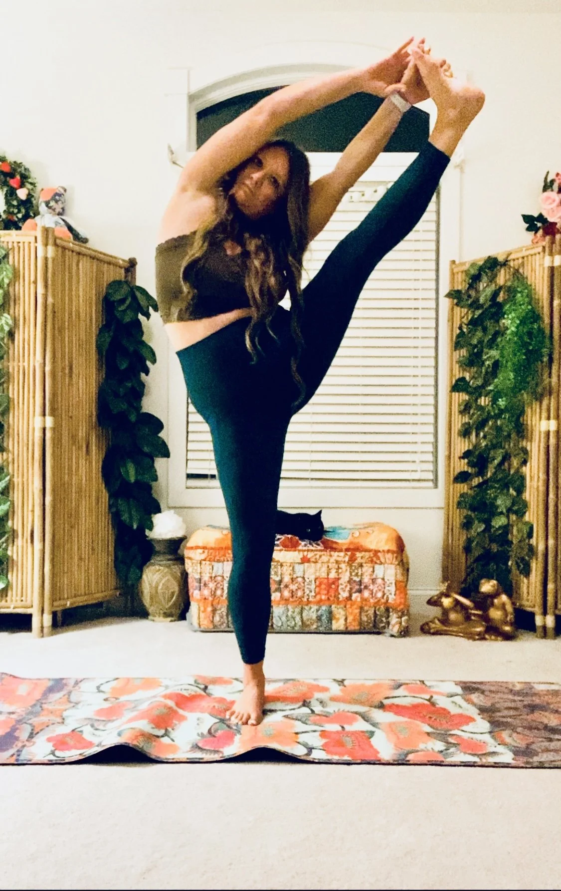 Woman practicing yoga indoors, performing a standing split pose on a floral yoga mat, surrounded by plants and decorative items.