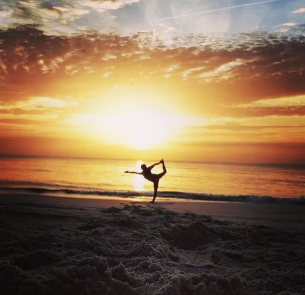 Person performing a yoga pose on the beach during sunset, with the sky full of colorful clouds and the ocean in the background.
