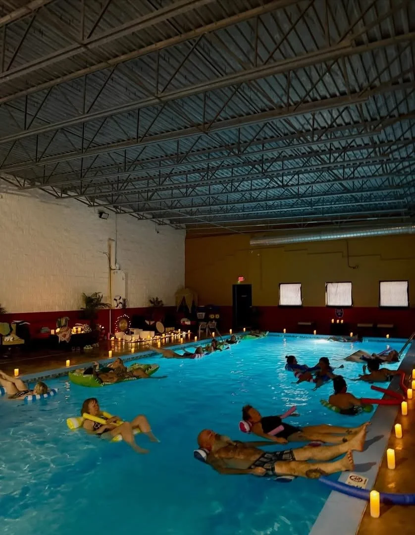 Indoor pool with people floating on pool noodles and relaxing, surrounded by small candles along the pool’s edge, with a high ceiling and windows.