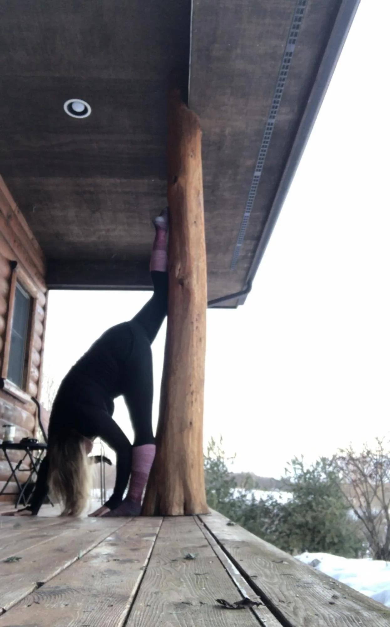A person standing upside down on a wooden deck, leaning against a large wooden pole, with a log cabin in the background and snow on the ground.
