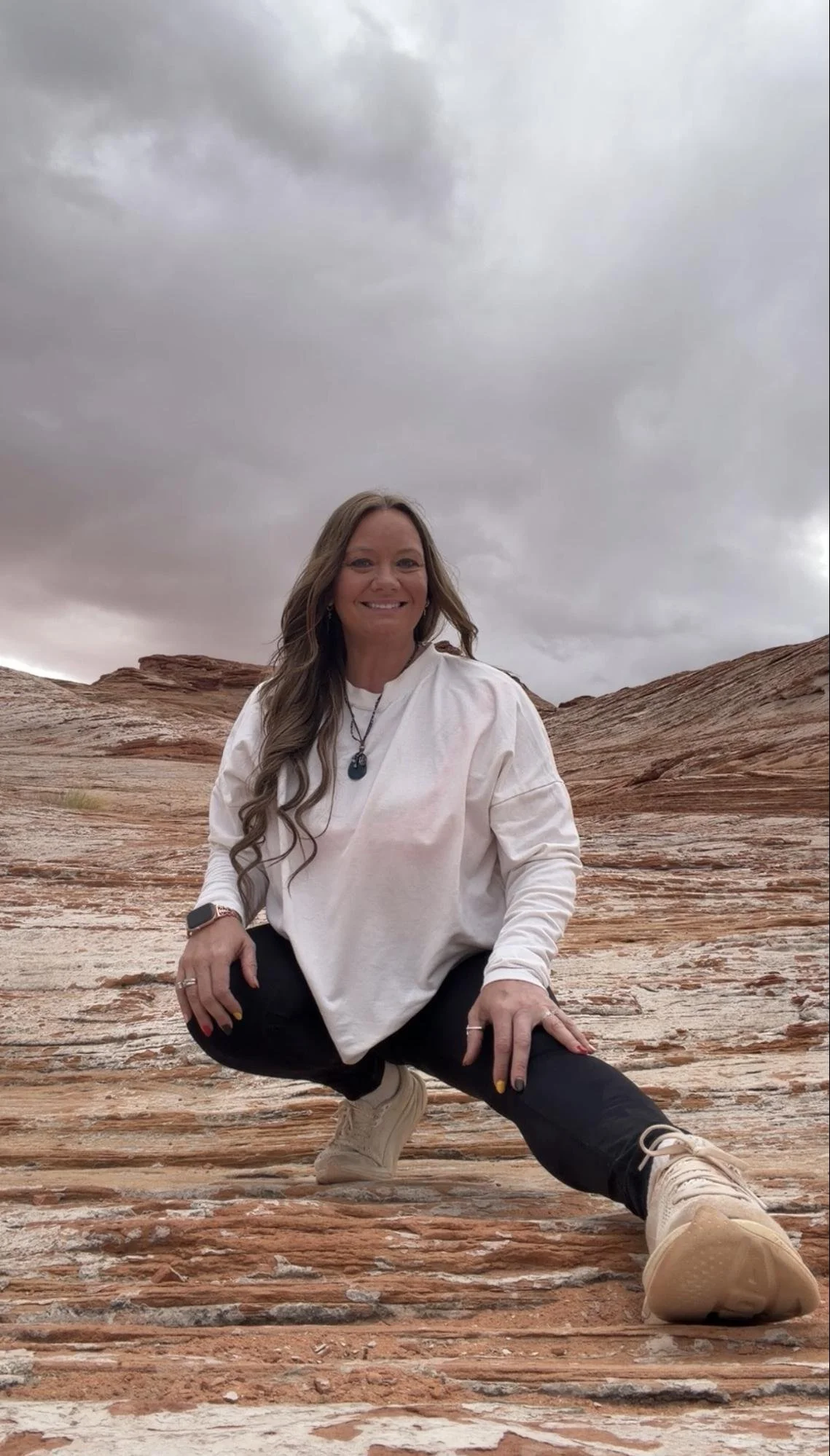 A woman squatting outdoors in front of layered red rocks under a cloudy sky, smiling at the camera.