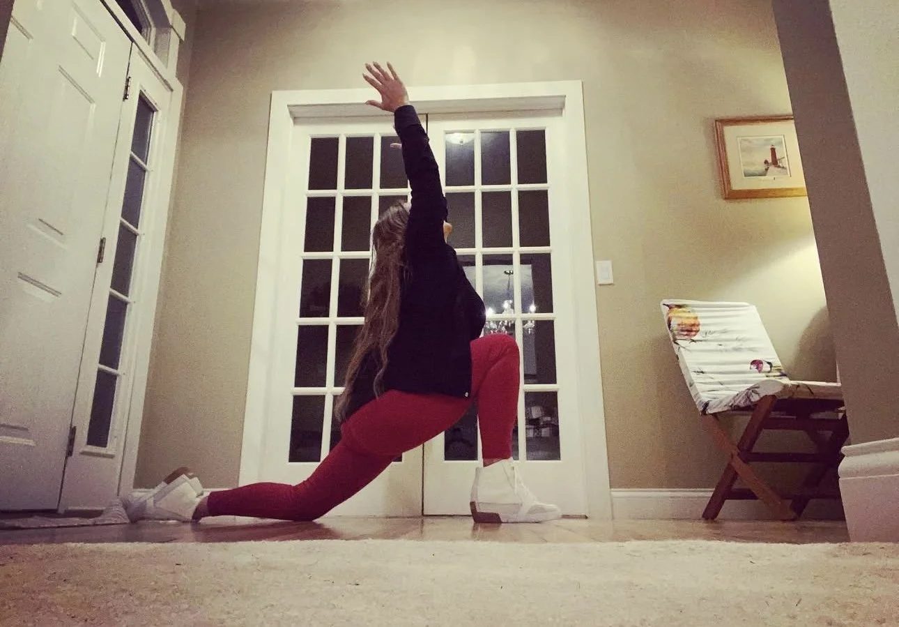 A woman performing a lunge stretch in a living room, with her right knee on the floor and her left knee bent at a 90-degree angle, reaching her left arm upward.