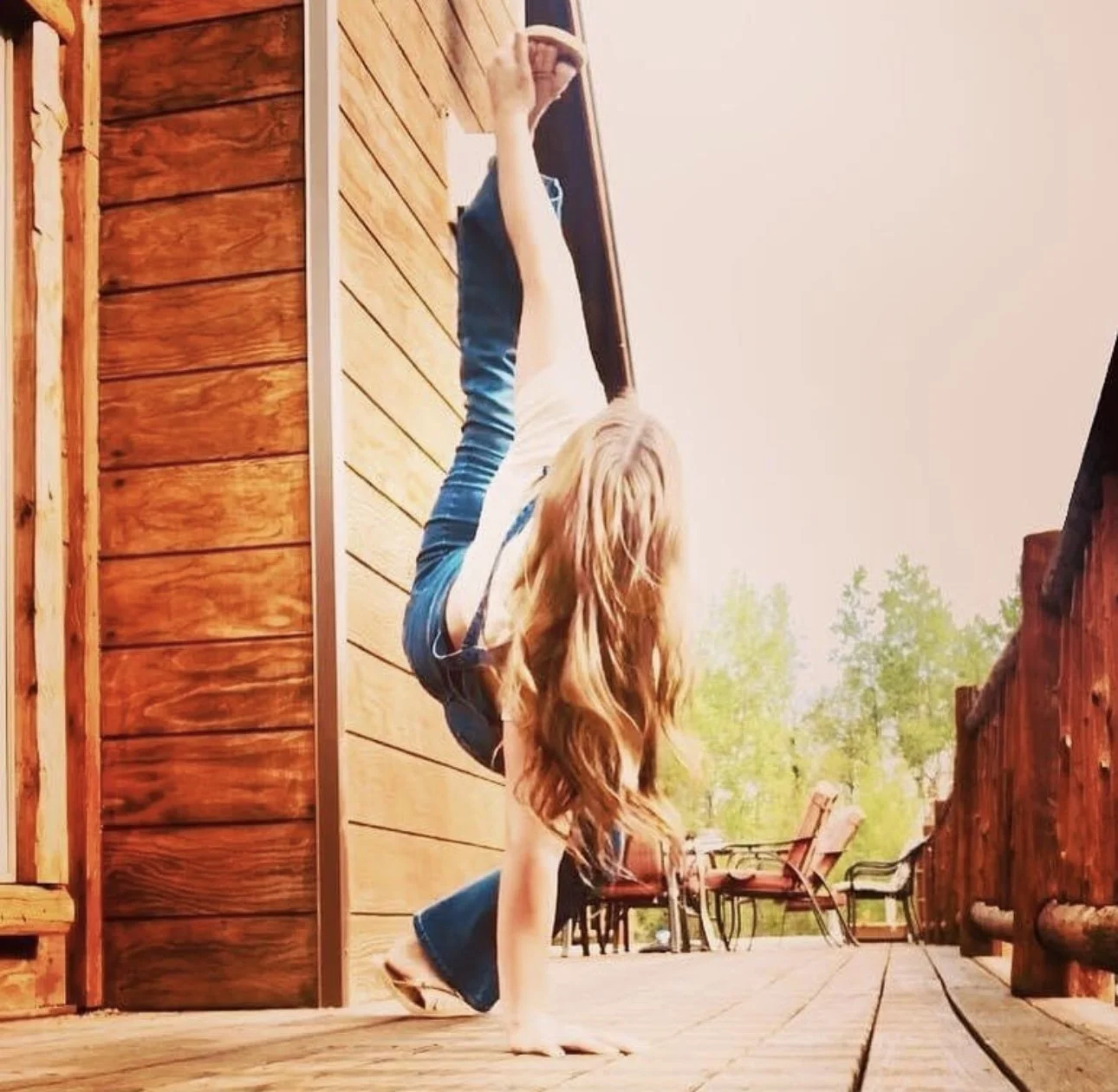 Young woman with long wavy hair performing a handstand on a wooden deck, with a backdrop of outdoor chairs and trees.