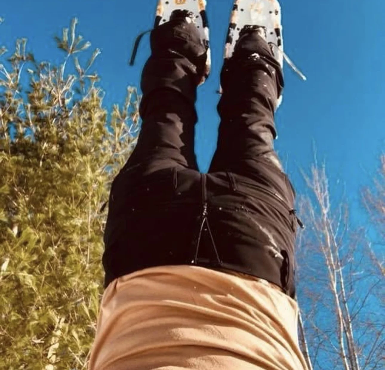 Person standing outdoors, wearing beige pants and a black jacket, with trees and a blue sky in the background.